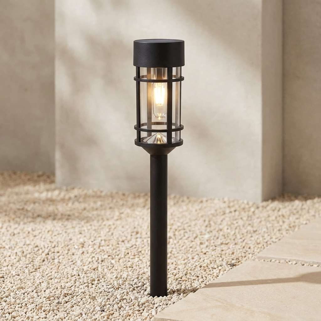 A black outdoor landscape light with a cylindrical glass shade and warm glowing bulb, set in gravel beside a stone path against a neutral concrete wall.