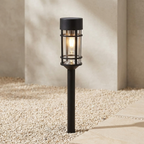 A black outdoor landscape light with a cylindrical glass shade and warm glowing bulb, set in gravel beside a stone path against a neutral concrete wall.