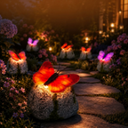 A garden pathway at night with glowing butterfly-shaped solar lights mounted on stone bases, surrounded by flowers and soft warm ambient lighting.
