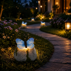 A garden pathway at night illuminated by butterfly-shaped solar lights placed on small rocks beside grass and flowers.