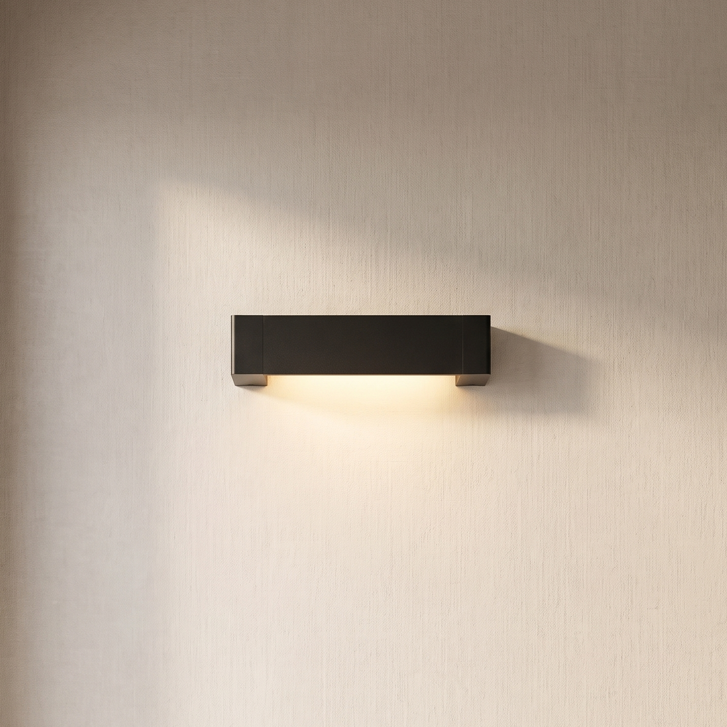 A minimalist rectangular wall sconce in matte black, mounted on a textured beige wall and casting a warm light downward, shown in a clean interior setting.