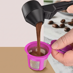 A hand holds a black coffee dripper above a bright pink reusable coffee filter cup, pouring coffee into it. A few coffee beans sit on a marble surface in the background.