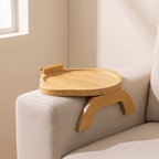 A light natural wood lap desk tray with curved legs and a small side hook, shown resting on the arm of a light beige sofa in a softly lit room.