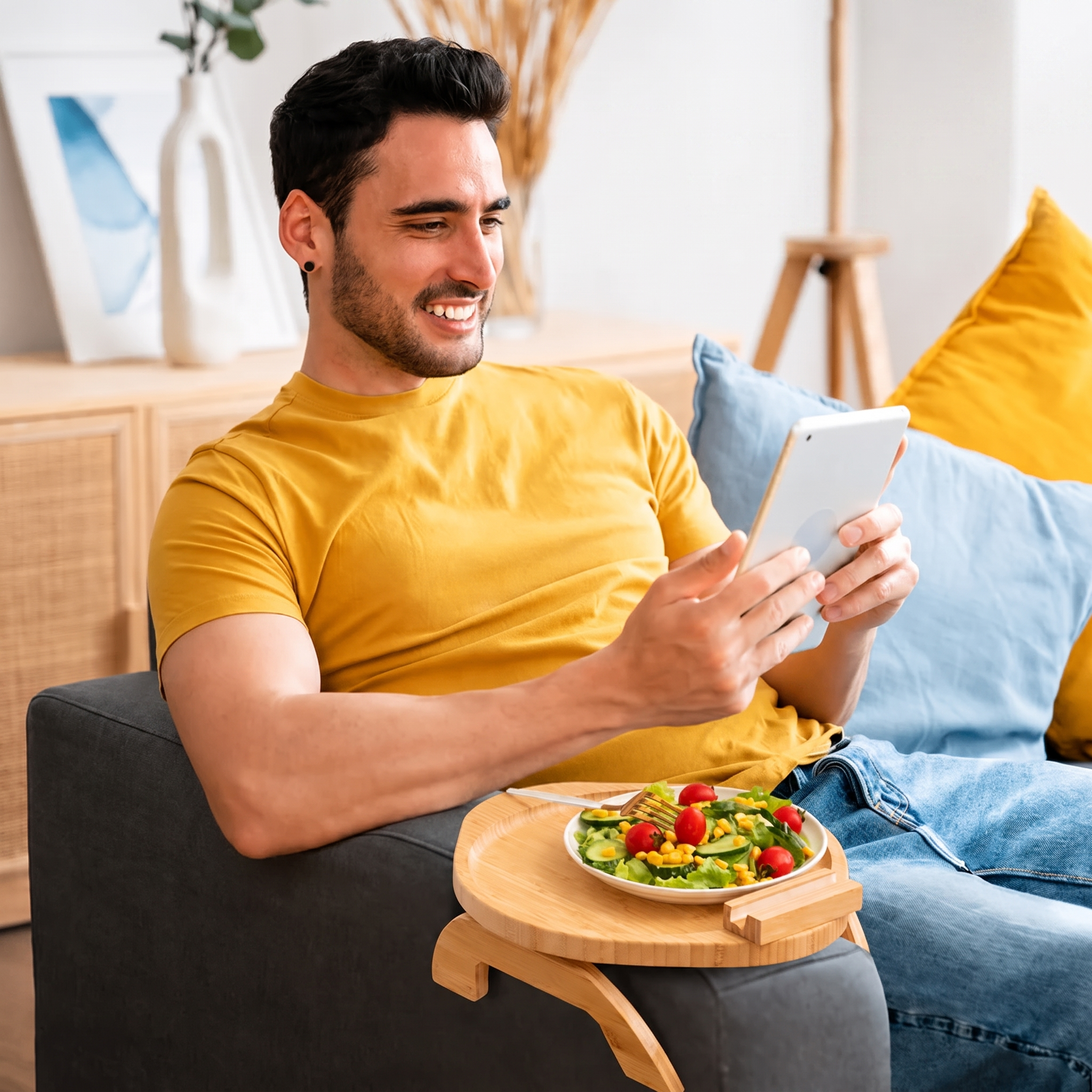 A man in a yellow t-shirt relaxes on a gray sofa, holding a white tablet and looking at it. A small wooden tray holding a plate of salad sits on his lap.