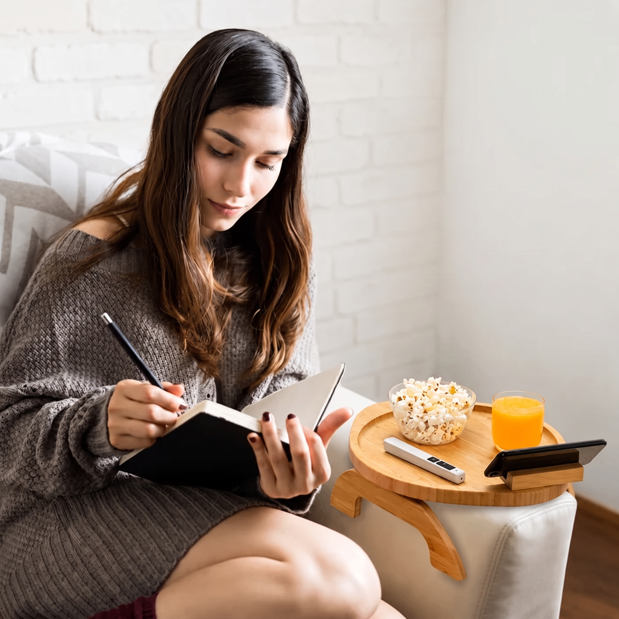 A woman in a brown knit sweater sits on a white armchair, writing in a notebook beside a small wooden side table holding a bowl of popcorn, a glass of orange juice, and a smartphone.