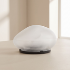 A modern, minimalist dome-shaped table lamp with a frosted white glass shade and a matte black circular base, set on a light beige surface against a softly blurred warm-toned background.