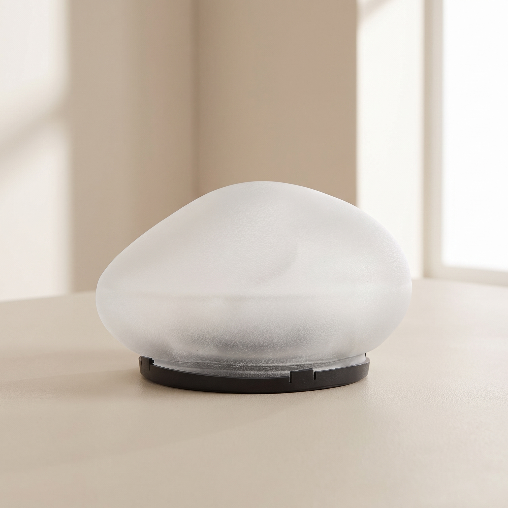 A modern, minimalist dome-shaped table lamp with a frosted white glass shade and a matte black circular base, set on a light beige surface against a softly blurred warm-toned background.
