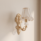 An ornate gold wall sconce with a textured clear glass shade and a clear crystal accent, mounted on a beige wall.