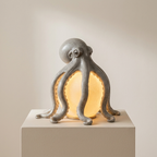 An octopus-shaped table lamp with a textured gray ceramic body and a glowing warm yellow light inside, displayed on a matching light gray pedestal against a neutral beige background.