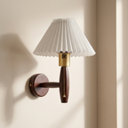 A wall-mounted sconce with a white pleated shade, a dark brown wooden stem, and brass metal accents, shown lit against a warm beige wall.