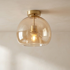 A modern semi-flush mount ceiling light with a large, rounded amber-tinted glass shade and a brushed brass metal fixture, shown illuminated against a neutral off-white ceiling.