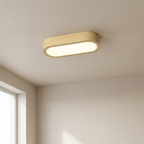 A modern oval ceiling light fixture with a light natural wood frame and a glowing white frosted glass shade, installed on a beige ceiling in a minimalist room.