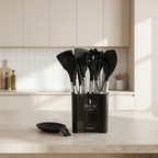 Black kitchen utensil set in a matching holder on a light countertop, with a matching black spatula placed beside the holder. The background shows a softly lit modern kitchen with cream cabinets and white tile backsplash.