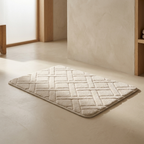 A rectangular bath mat with a quilted basketweave pattern in a warm off-white/cream color, shown on a matching beige floor in a minimalist bathroom setting.