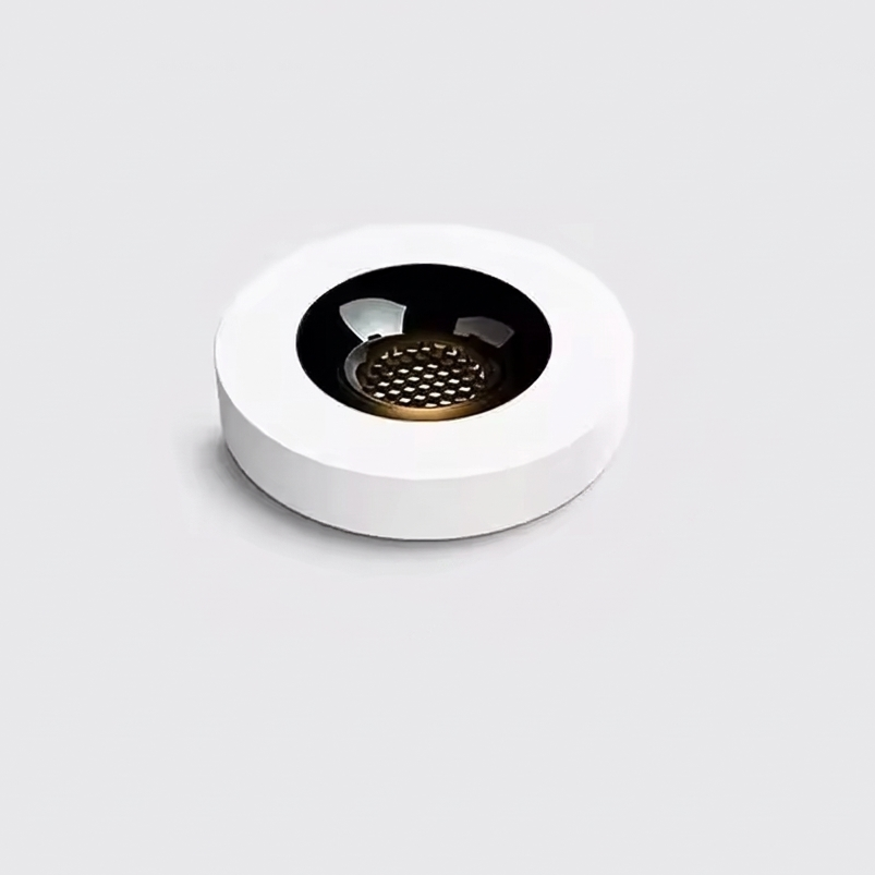 A minimalist white circular coffee cup holder or coaster with a black center and a gold-toned perforated metal insert, shown on a light gray background.