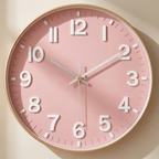 A round wall clock with a soft pink face, large white Arabic numerals, white hands, and a thin gold-toned outer rim, shown mounted on a light beige wall.