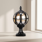 A black metal globe-style table lamp with a warm amber-tinted glass shade and a square pedestal base, shown on a light beige surface near a window.