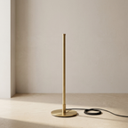 A minimalist table lamp with a slim vertical stem and a round circular base, finished in brushed brass/gold, shown on a light beige floor against a neutral beige wall with a black power cord.
