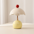 A small mushroom-shaped table lamp with a glossy pale yellow base, a cream-colored cap, a red spherical topper, and a metallic stem, shown on a light surface against a soft neutral background.