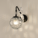 A wall-mounted light fixture with a black curved arm, a white sculptural figure, and a textured clear glass globe shade, shown lit against a warm off-white wall.