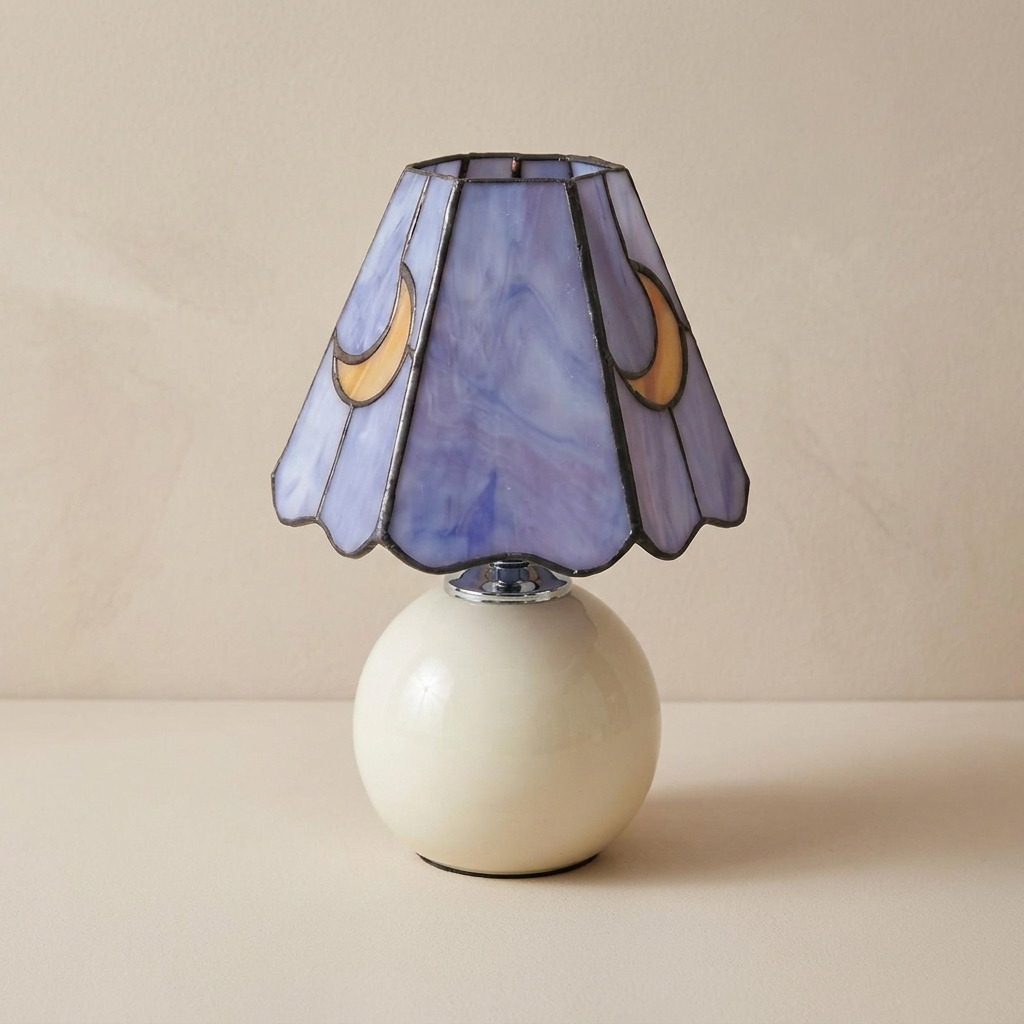 A table lamp with a sculptural, rounded off-white ceramic base and a stained-glass style conical shade in lavender and blue with gold crescent moon motifs, set against a neutral beige background.