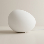 A minimalist white egg-shaped table lamp with a smooth matte finish, resting on a light beige surface against a matching neutral background.