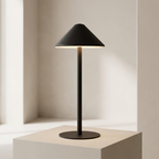 A modern black table lamp with a conical shade and cylindrical base, illuminated and placed on a light beige pedestal against a minimalist beige background.