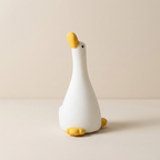 A white duck-shaped night light with a yellow beak, feet, and eye, standing on a light beige surface against a matching beige background.
