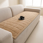 A beige quilted corduroy sofa pad on a light gray fabric sofa, with a small black cup and saucer placed on top, in a bright, modern living room setting.