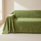 An olive green textured throw blanket with fringed edges draped over a sofa in a minimalist, sunlit room.