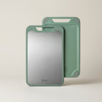 Two sage green plastic cutting boards with integrated handles, one showing a brushed silver metallic cutting surface and the other showing the solid green back, on a warm off-white background.