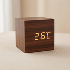 A square wooden digital clock with a dark brown wood grain finish and glowing yellow LED numbers showing 26°C, placed on a light beige fabric surface with a soft neutral background.