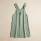 A sage green linen pinafore dress with a split neckline, wide shoulder straps, and two front patch pockets, laid flat against a warm beige background.