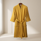 A mustard yellow waffle-knit bathrobe with a tie belt and front pockets, hanging on a wooden hanger against a warm off-white background.