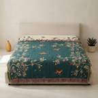 A teal woven throw blanket with a bird and floral pattern is styled on a bed with a beige upholstered headboard. The scene is set in a minimalist room with a small vase on the left and a potted succulent on the right.