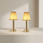 Two matching table lamps with pleated beige fabric shades and polished gold bases, displayed side by side on a white surface against a warm off-white wall.