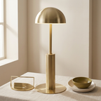 A modern table lamp with a dome-shaped shade and cylindrical base in brushed brass, styled on a light beige tablecloth next to a small bowl and tray in a softly lit room.