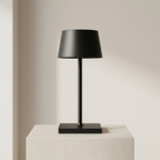 A matte black table lamp with a tapered drum shade, slim stem, and square base, displayed on a textured white pedestal against a warm beige wall.