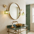 A bathroom vanity scene with a round wall mirror, two brass sconces with green glass shades, a brass vessel sink on a green marble console, and a tiled shower area in the background.