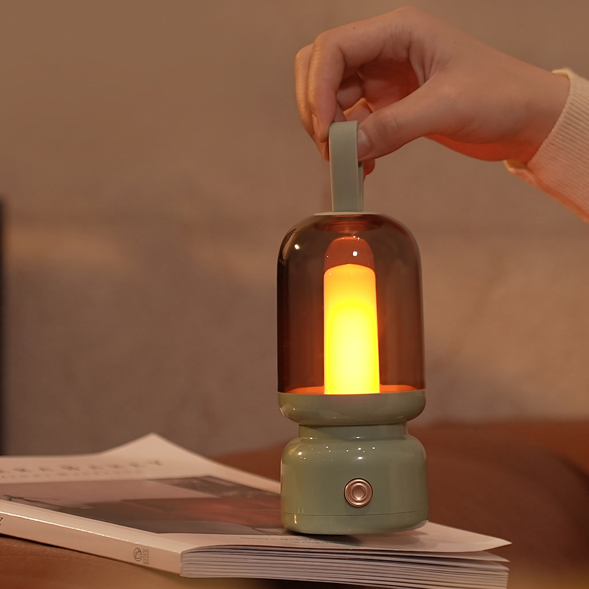 A hand holds a small lantern-style lamp with a glowing warm light inside a translucent smoky dome, resting on an open book in a softly lit indoor setting.