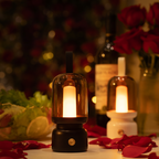 A lit amber glass lantern with a black base sits on a table surrounded by rose petals, with a second candle and blurred flowers in the background.