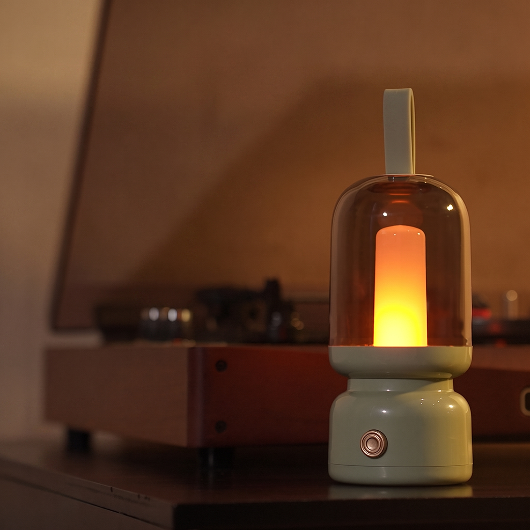A small tabletop lantern with a translucent amber glass dome and a glowing warm light inside, set on a dark surface in a dim room with a blurred record player in the background.