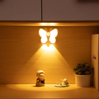 A butterfly-shaped LED night light glowing with warm yellow light on a wooden shelf, with small decorative figurines and a potted plant below.