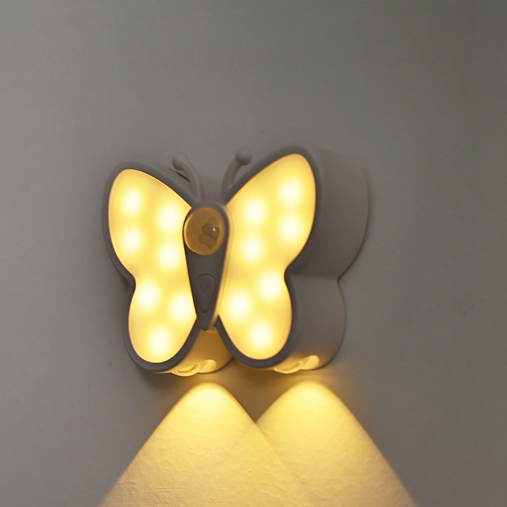 A butterfly-shaped wall night light with glowing warm yellow LED lights, mounted on a plain gray wall and casting a soft downward light.