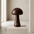 A dark brown wooden mushroom-shaped table lamp with a small gold metal accent, displayed on a textured beige pedestal in a minimalist, warm-toned interior setting.