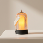 A ribbed glass lantern-style table lamp with a warm glowing light, a white handle, and a black base, photographed on a clean white tabletop against a soft beige wall.