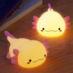 Two cute axolotl-shaped night lights glowing with warm yellow light on a wooden surface, with a blurred book in the background.