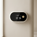 A modern oval digital clock or alarm unit with a black display screen and a gold button, mounted on a beige wall.