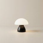 A mushroom-shaped table lamp with a glowing white dome shade and a matte black base, set against a minimalist beige background.