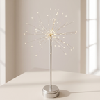 A table lamp with a silver metal stand and circular base, featuring a starburst-style wire frame filled with warm white LED lights, set on a light-colored surface against a softly lit neutral background.